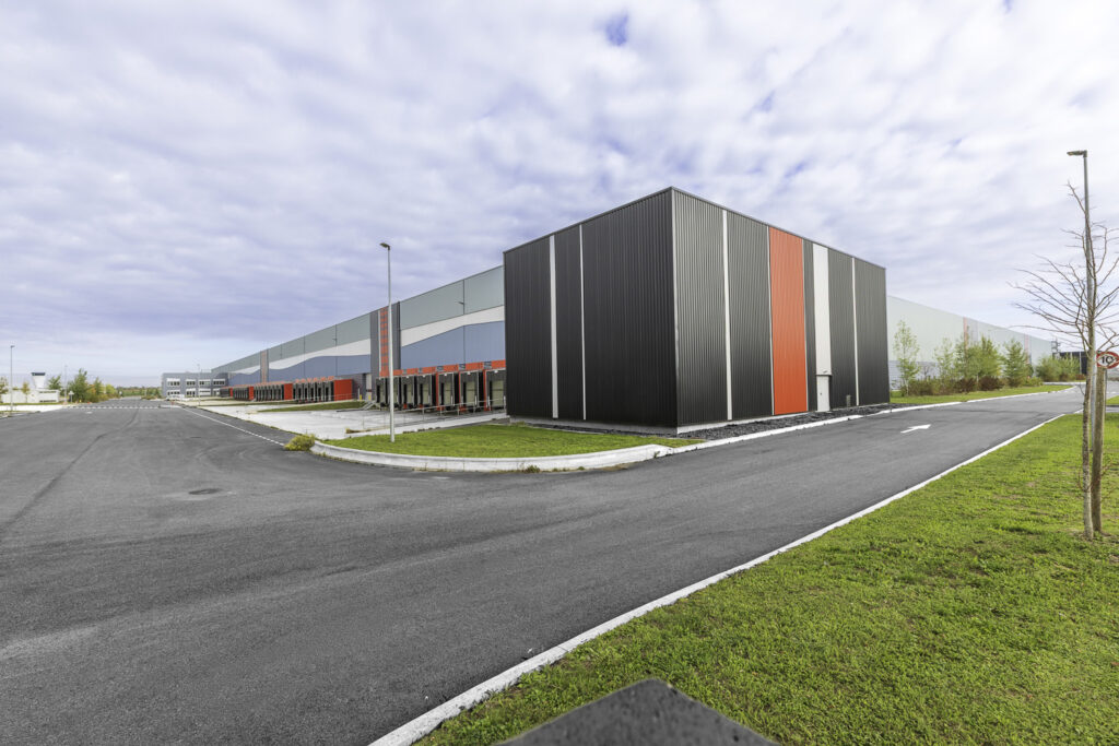 Vue extérieure du futur centre de distribution Amazon à Novaparc, à Beauvais, un vaste bâtiment logistique moderne entouré de voiries et d’espaces verts.